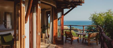 Una terraza de madera con vista al océano, una zona de estar con una mesa y sillas, y un ventanal que da a una habitación.