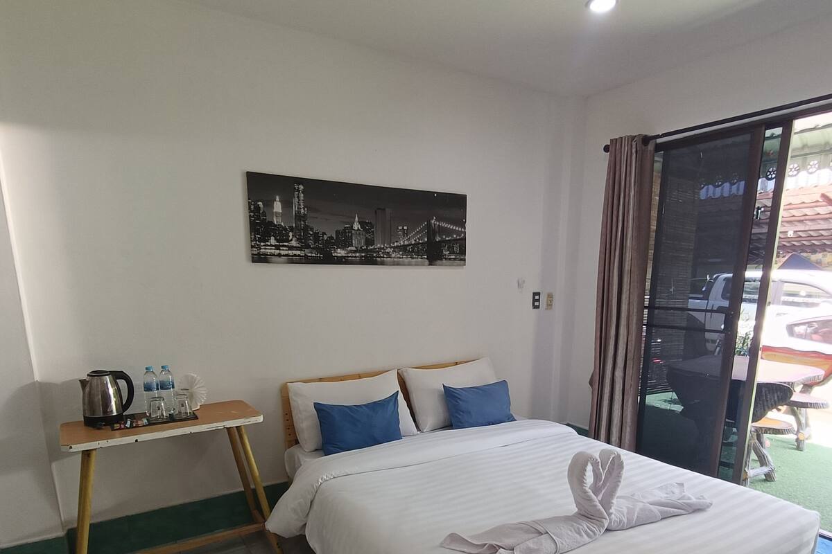 A hotel room with a bed, a small table with a kettle and water bottles, a cityscape painting on the wall, and a sliding glass door to the outside.