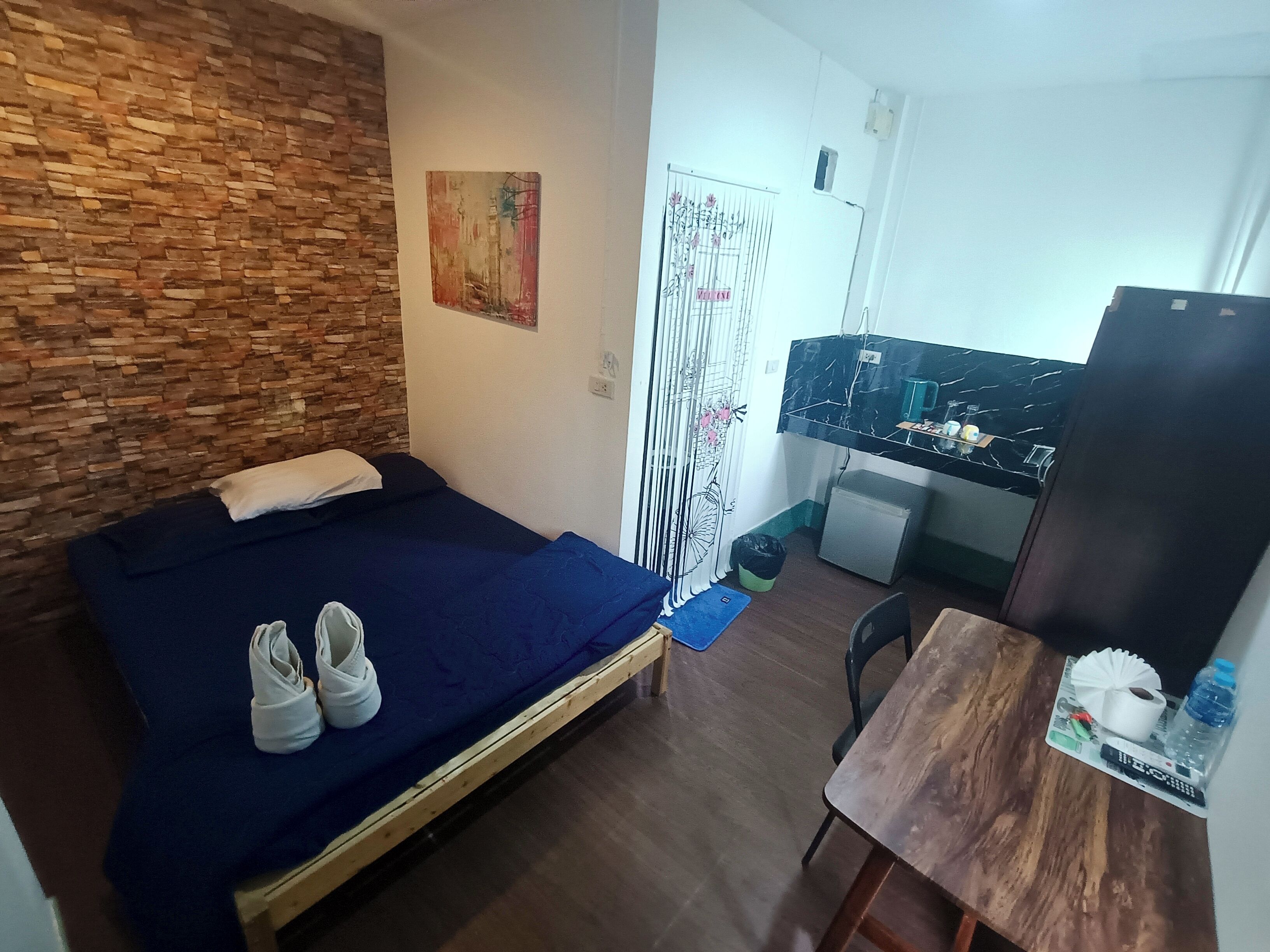 A bedroom with a wooden wall, a bed with a blue bedspread, a desk with a computer, and a small fridge.