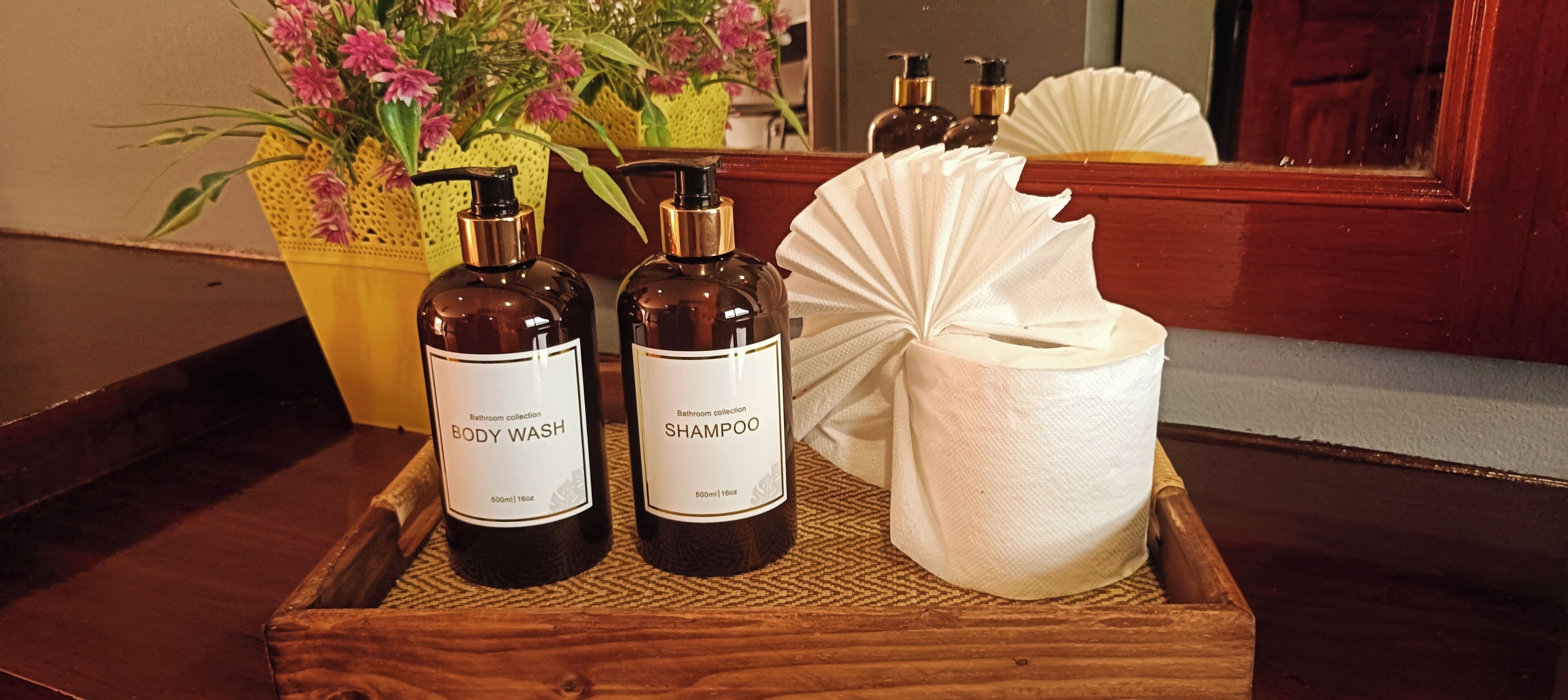 A wooden tray with two bottles of body wash and shampoo, a white paper towel, and a potted plant with pink flowers.