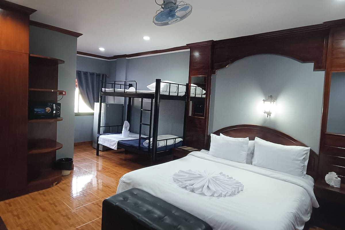 A hotel room with a bunk bed, a single bed, a wooden bench, a ceiling fan, and a wall-mounted light fixture.