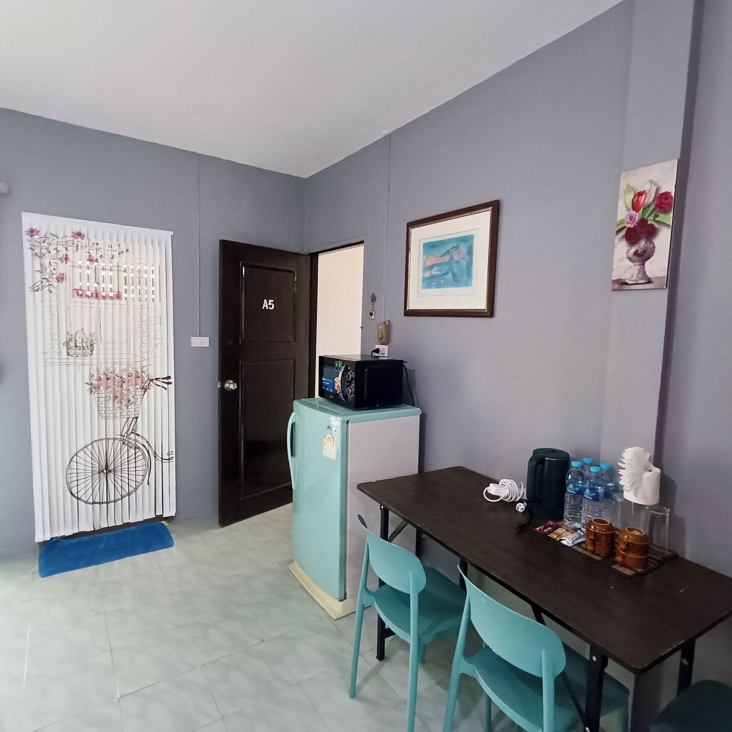 A compact room with a kitchenette, a dining table with chairs, a refrigerator, and a door marked A5.