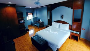 A hotel room with a bed, a desk, a chair, a small table, a mirror, and a ceiling fan. - The Guest House  (Patong)
