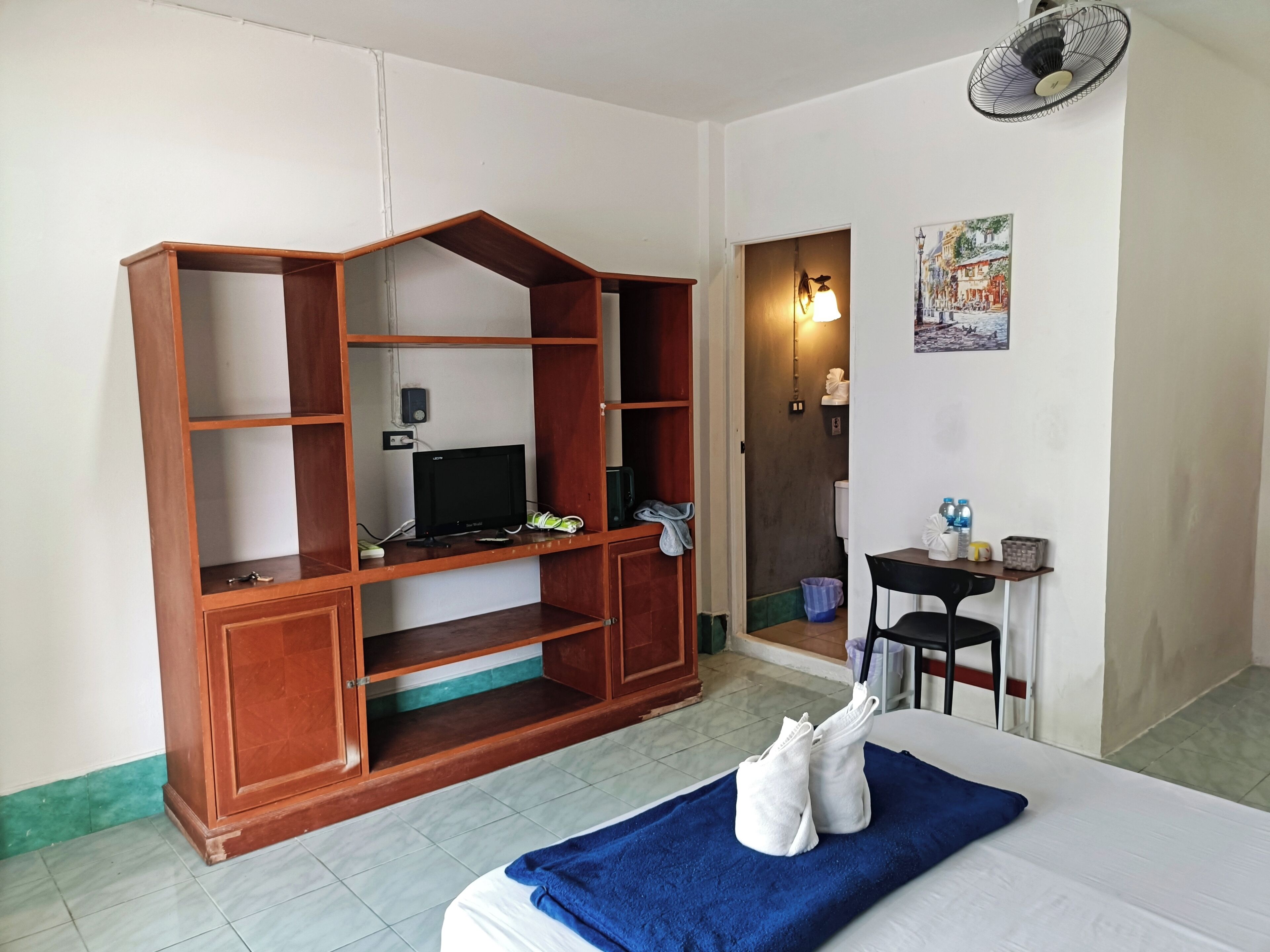 A hotel room with a wooden TV stand, a bed with a blue towel, a small table with a chair, and a fan.
