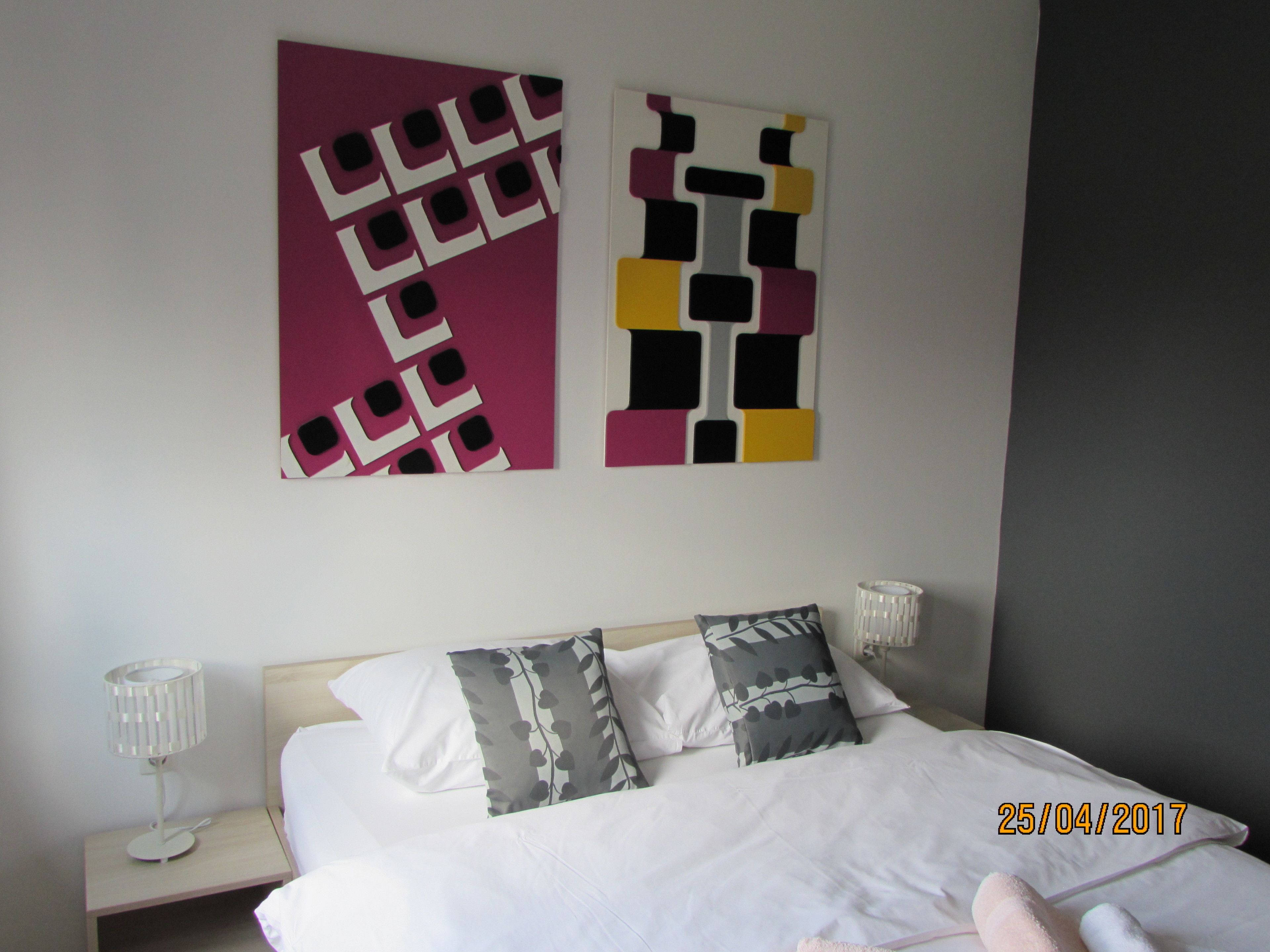 A hotel room with a bed, two pillows, a nightstand with a lamp, and two wall art pieces.