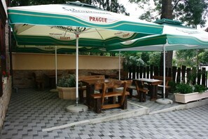An outdoor seating area with wooden tables and chairs under striped umbrellas, a brick wall, and potted plants.