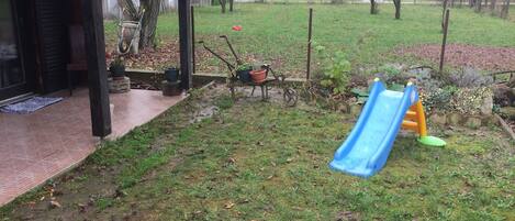 Een achtertuin met een blauwe glijbaan, een houten schommel en een terras van bakstenen.