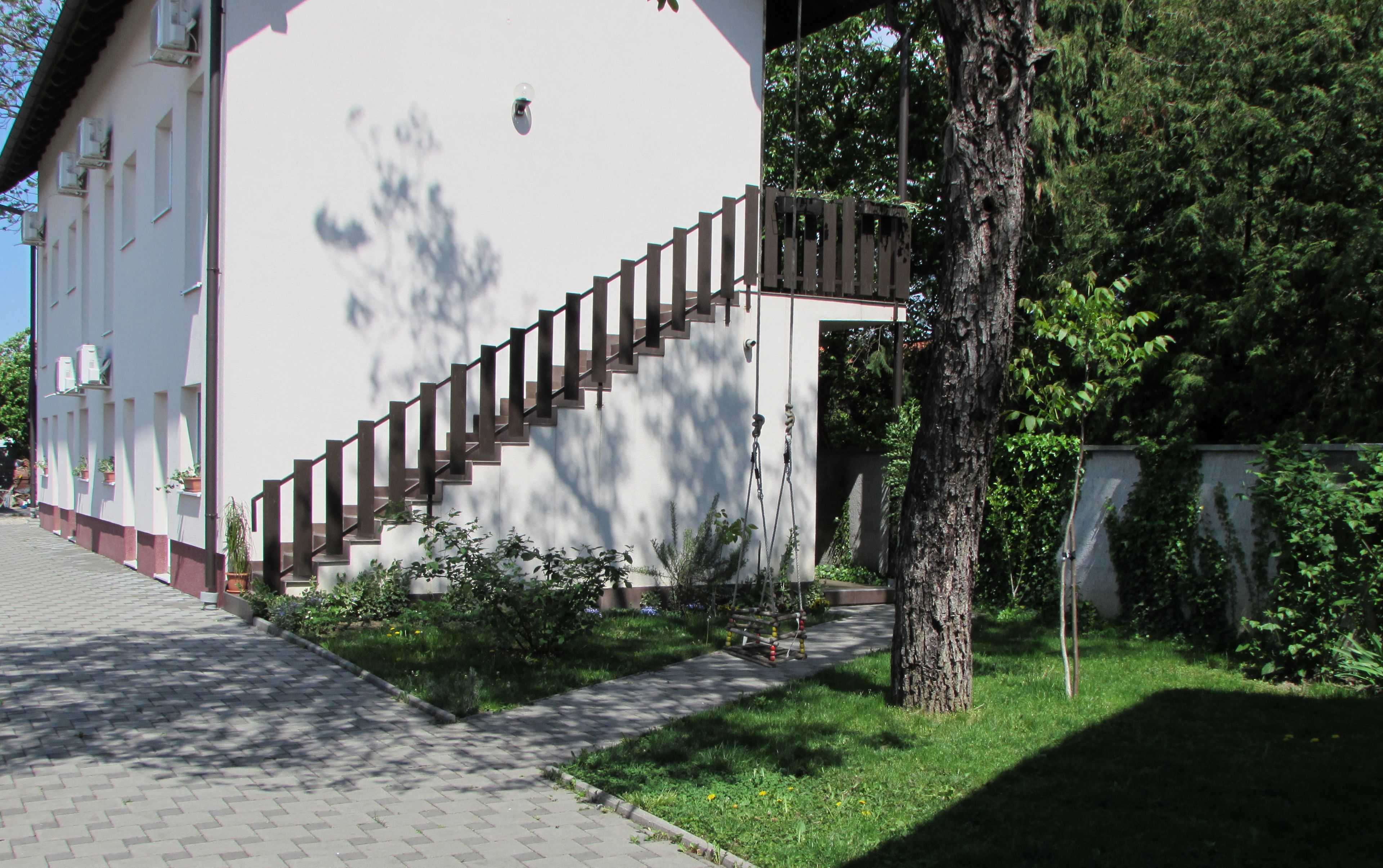 A white building with a staircase leading to a door, surrounded by greenery and a paved walkway.