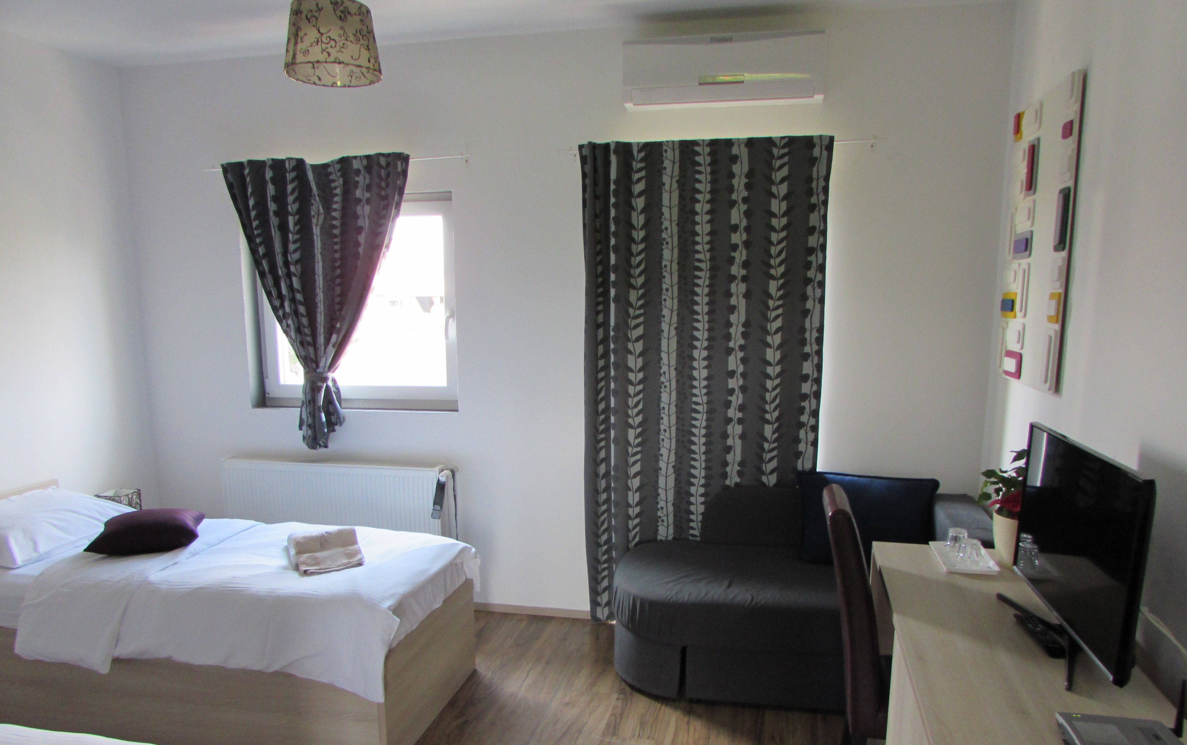 A bedroom with a bed, a chair, a desk with a computer, and a window with curtains.
