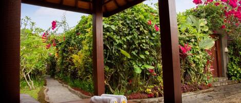 A wooden gazebo with a bench and a towel, surrounded by lush greenery and vibrant flowers.