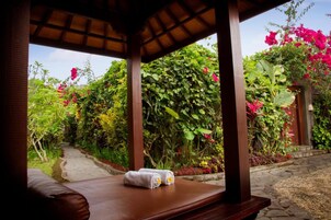 A wooden gazebo with a bench and a towel, surrounded by lush greenery and vibrant flowers.