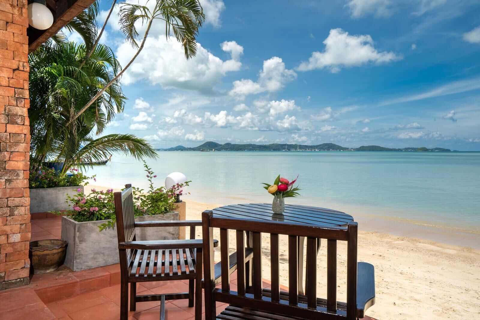 Ein Holztisch und -stuhl-Set auf einer Terrasse mit Blick auf einen Strand, Palmen und einen klaren Himmel.