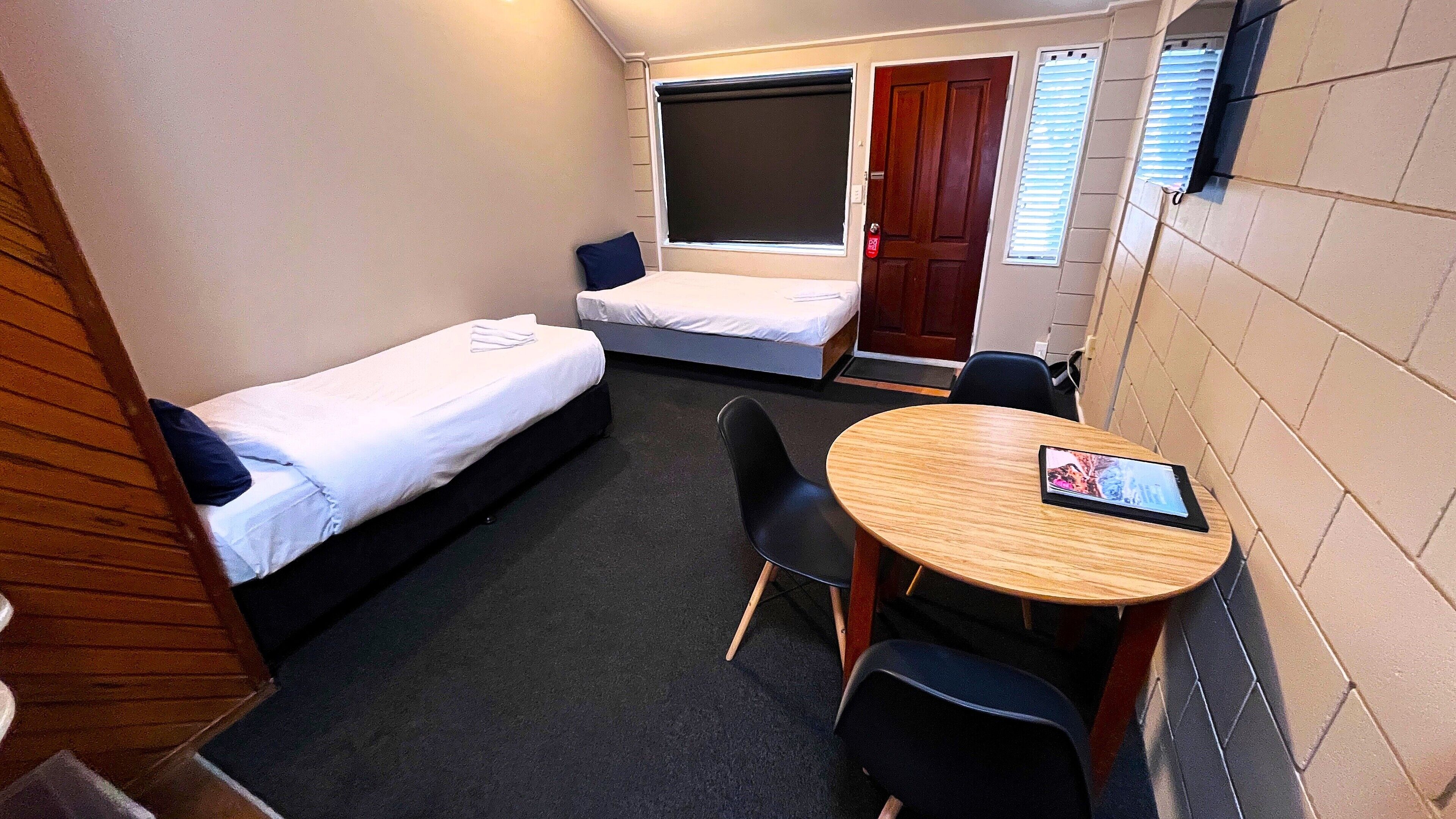Une chambre d’hôtel de taille modeste, équipée de deux lits simples, d’une petite table avec une chaise et d’une fenêtre avec des stores.
