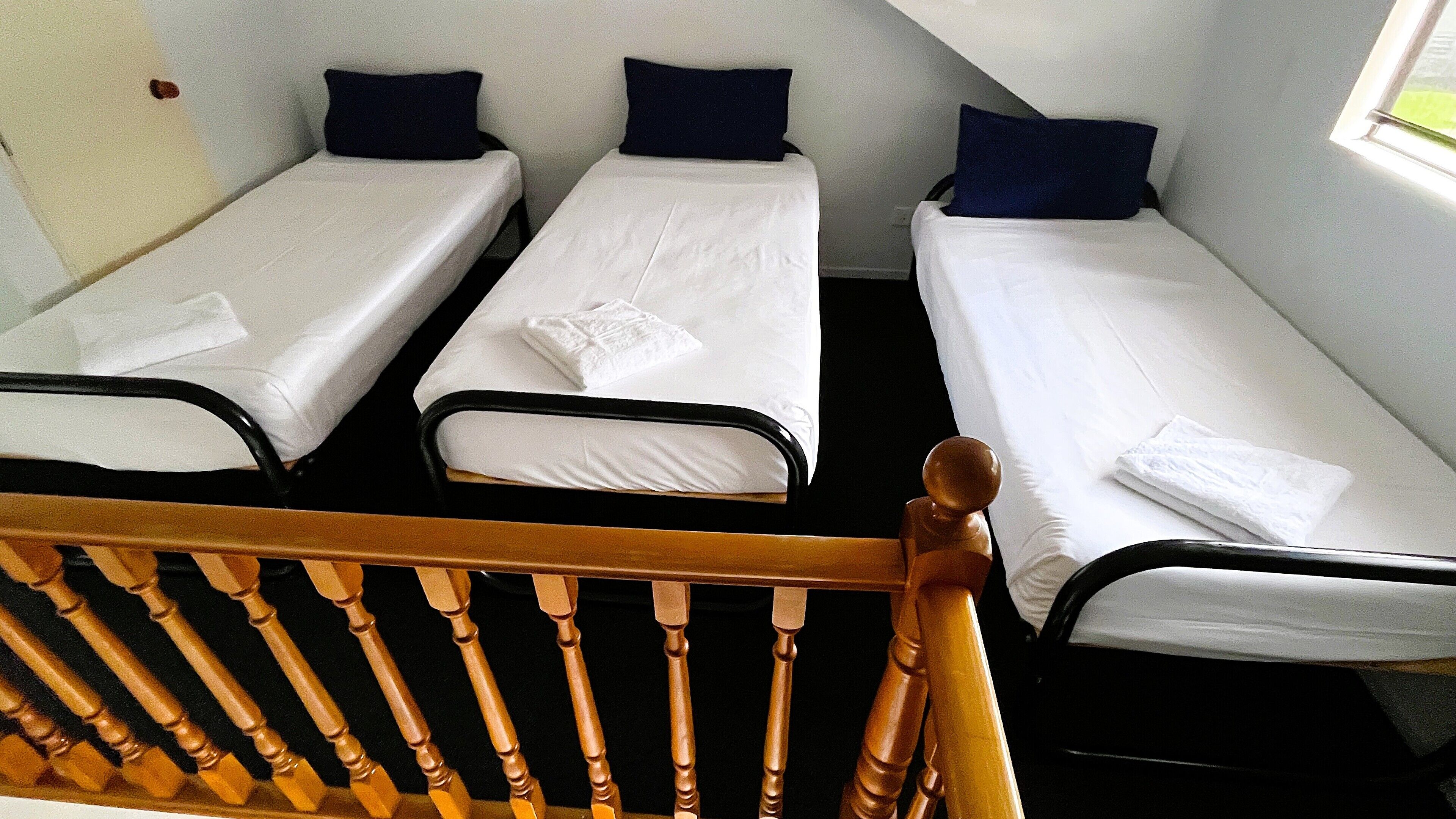 A room with three single beds, each with a pillow and a blanket. There is a wooden chair in the foreground.