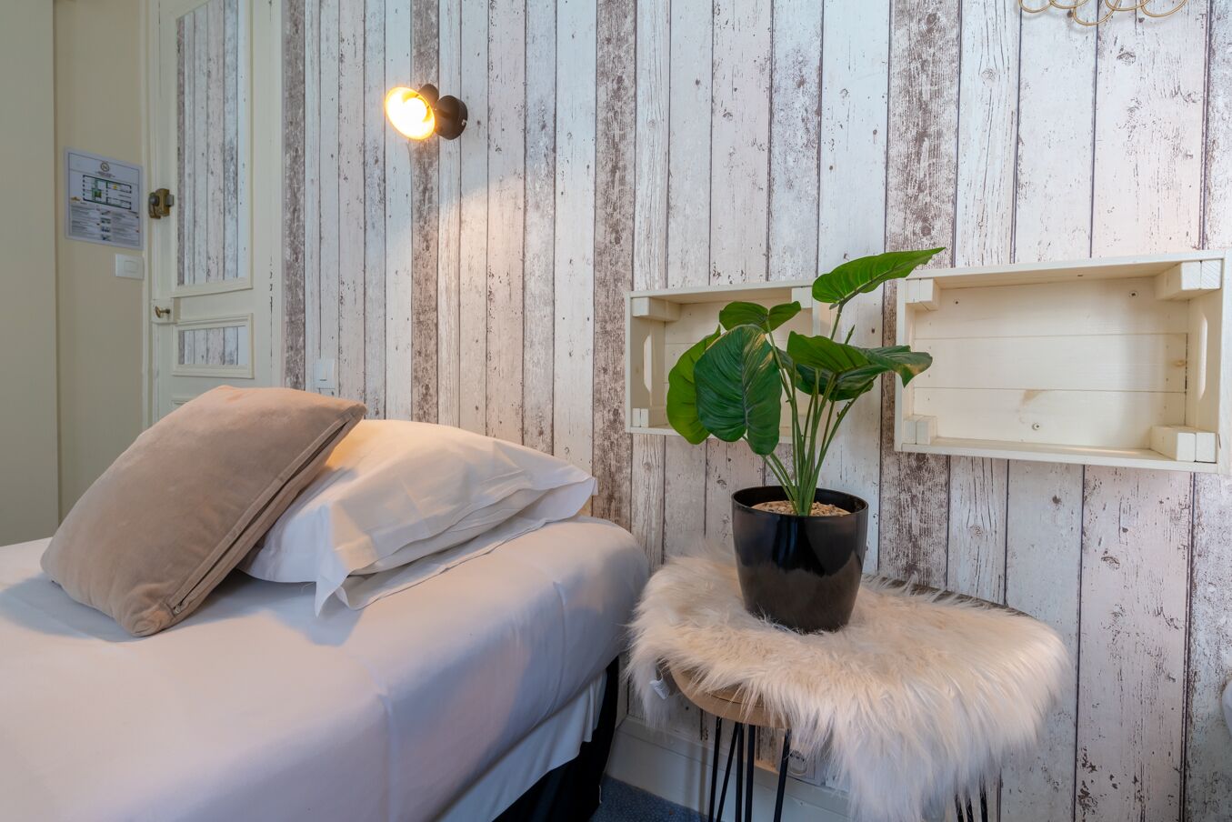 A bedroom with a bed, a nightstand with a plant, a wall-mounted shelf, and a wooden wall with a door.