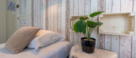 A bedroom with a bed, a nightstand with a plant, a wall-mounted shelf, and a wooden wall with a door.