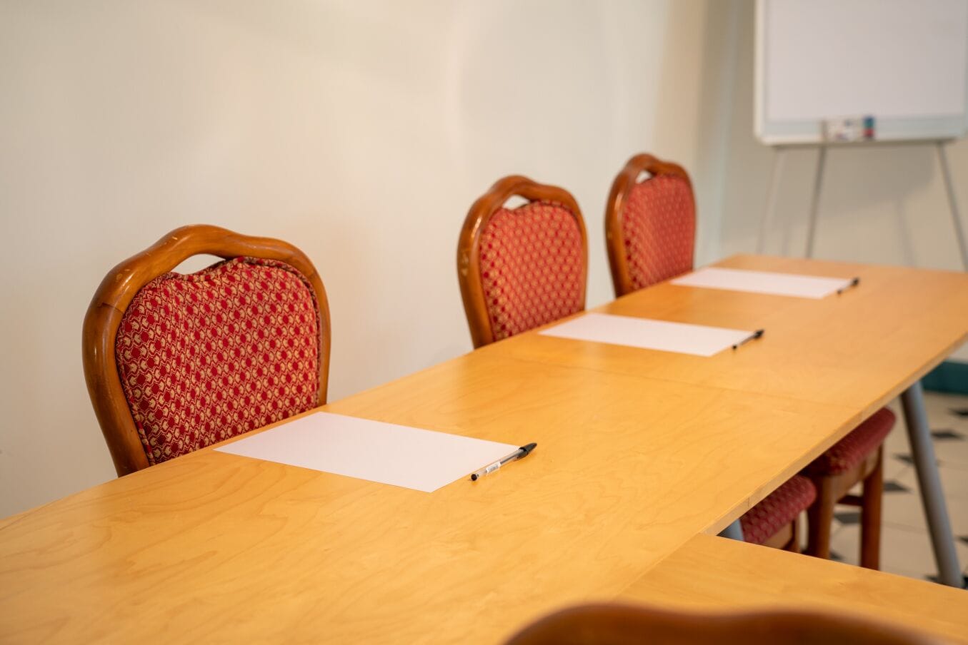 Ett konferensrum med ett långt bord i trä, fyra röda stolar med mönster och en whiteboard i bakgrunden.