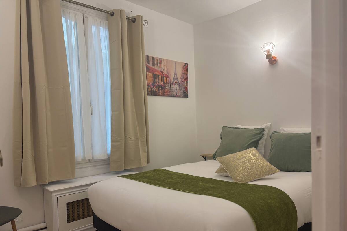Un dormitorio con cama, almohadas verdes y blancas, una manta verde, una ventana con cortinas, un radiador y una lámpara fijada a la pared.