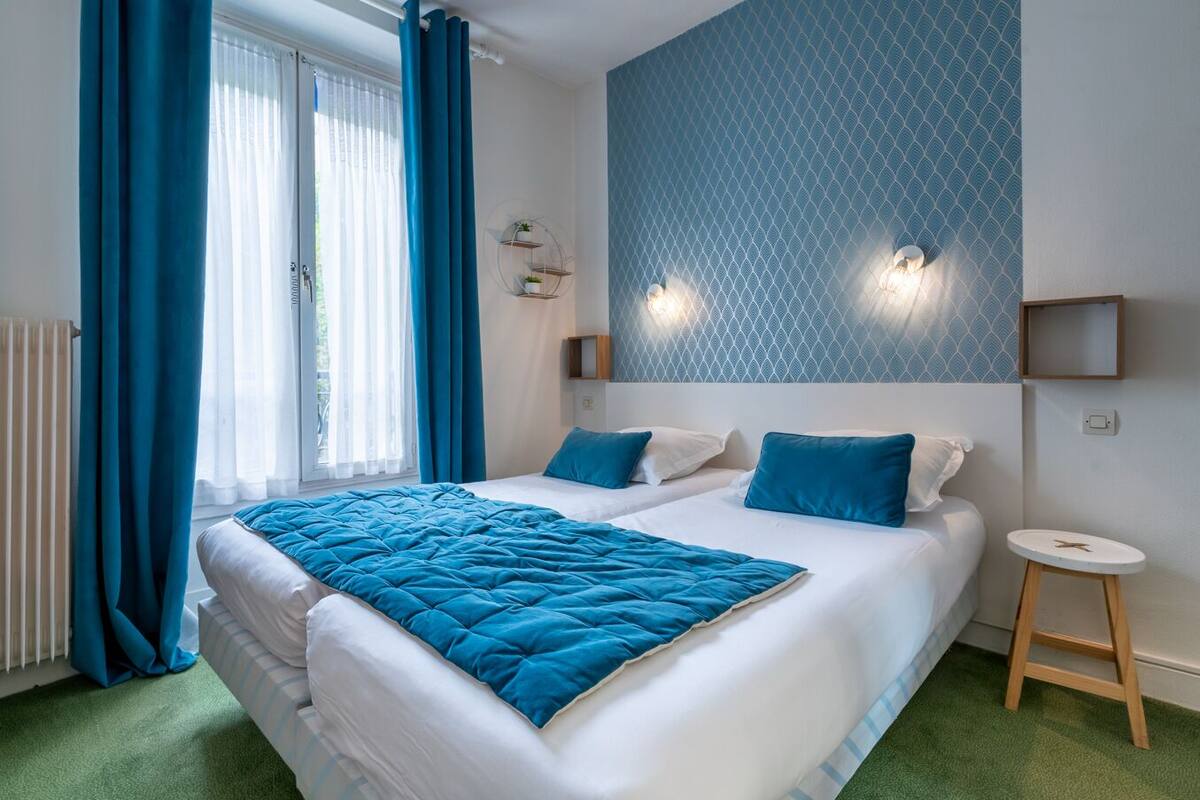 Habitación de hotel con una cama grande, dos mesitas de noche, un taburete pequeño, una lámpara de pared y una ventana con cortinas.