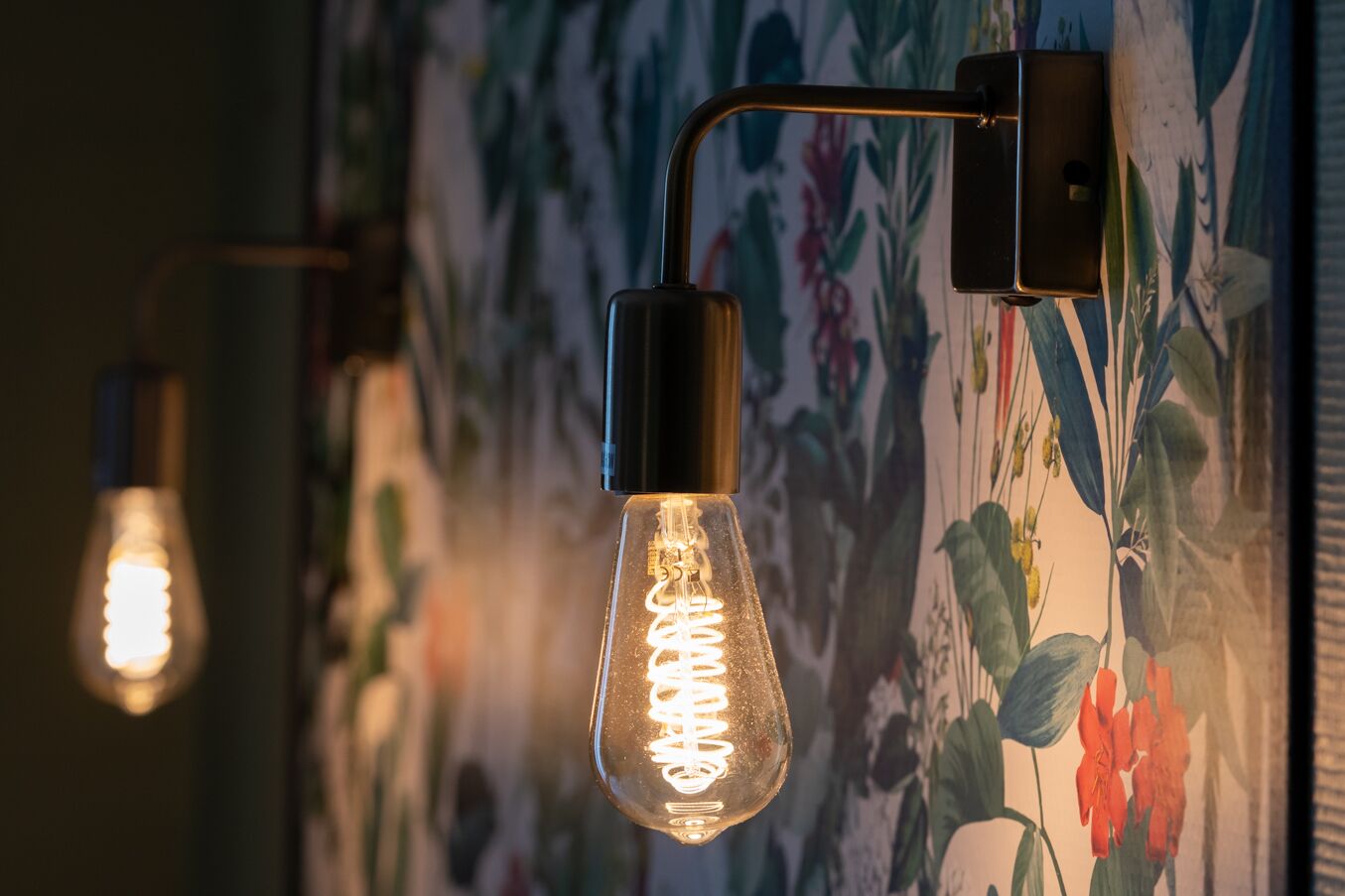 A wall-mounted light fixture with a clear glass bulb and a metal base, set against a floral wallpaper background.