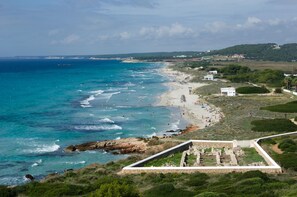 A coastal beach with clear blue waters, a sandy shore, and a stone structure in the foreground. - Royal Son Bou Family Club (Alayor)