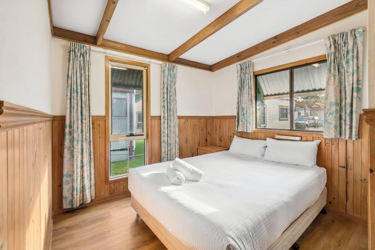 A wooden-paneled bedroom with a large bed, two windows with curtains, and a view of the outside.