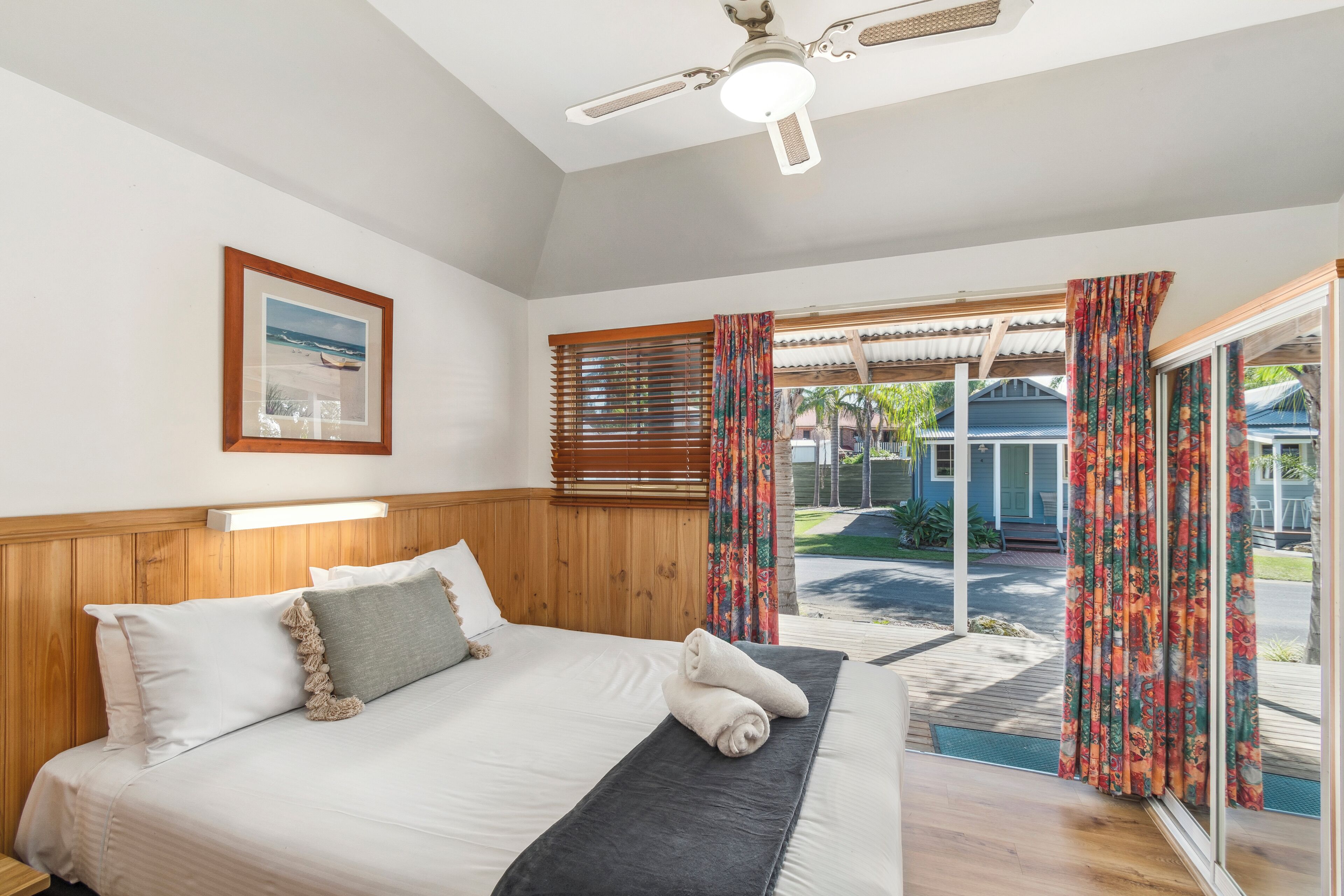 A bedroom with a bed, wooden headboard, a ceiling fan, a framed picture on the wall, and a view of the outdoors through a sliding glass door.