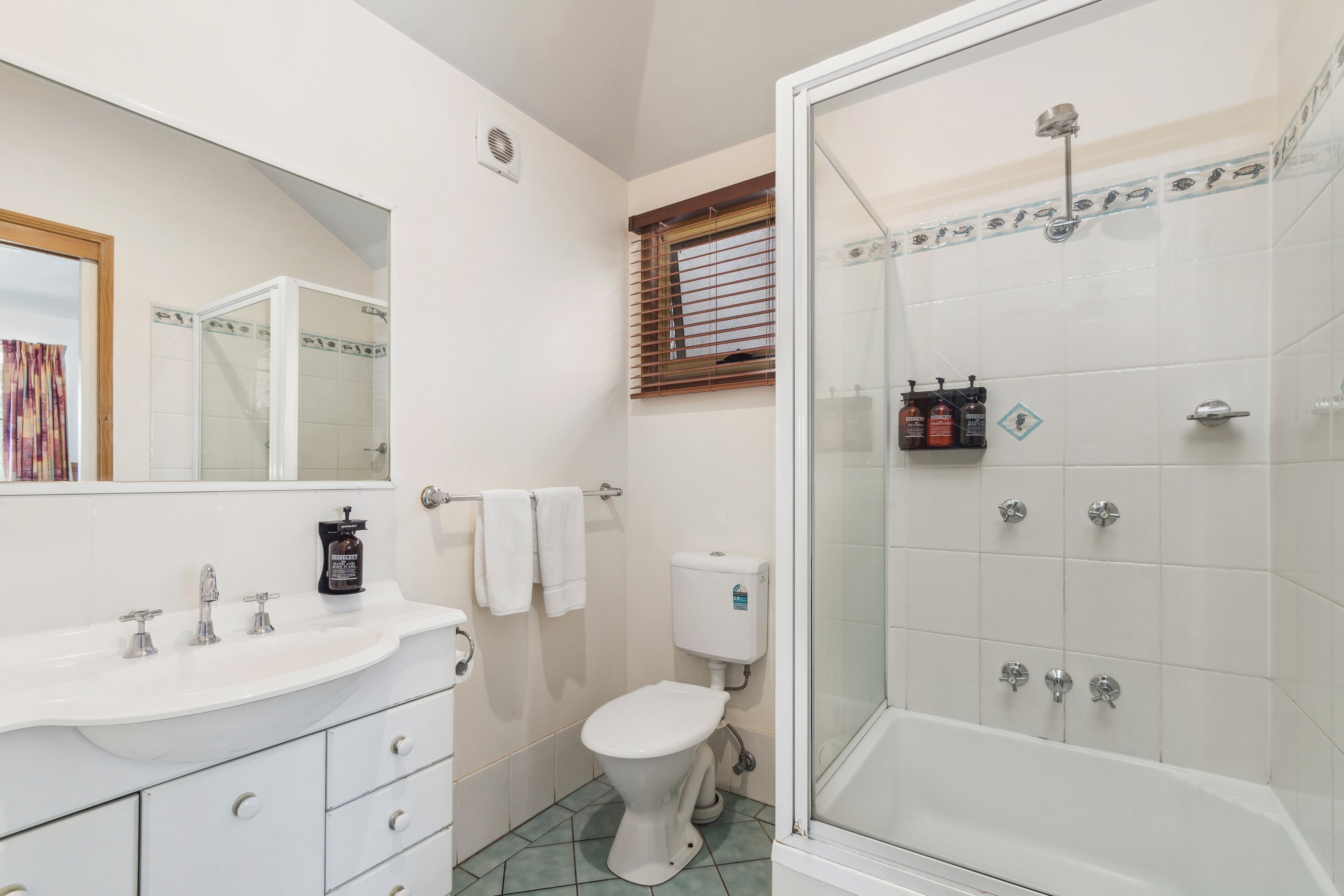 Een badkamer met een wit wastafel, toilet en douche. Handdoeken hangen aan een rek en er is een raam met zonwering.
