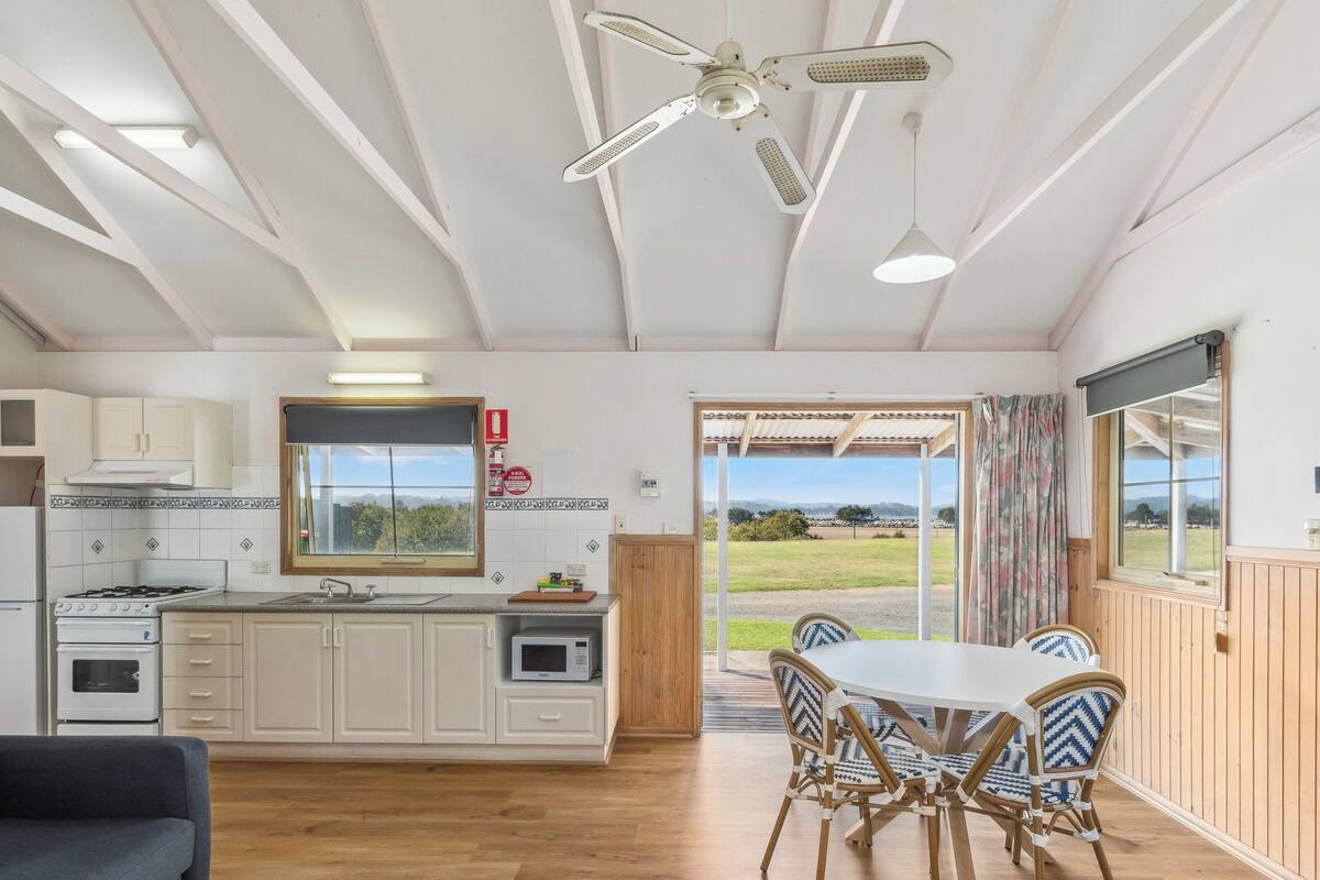 A spacious kitchen with white cabinets, a stove, and a microwave. There is a dining area with a table and chairs, and a large window offering a view of the outdoors.