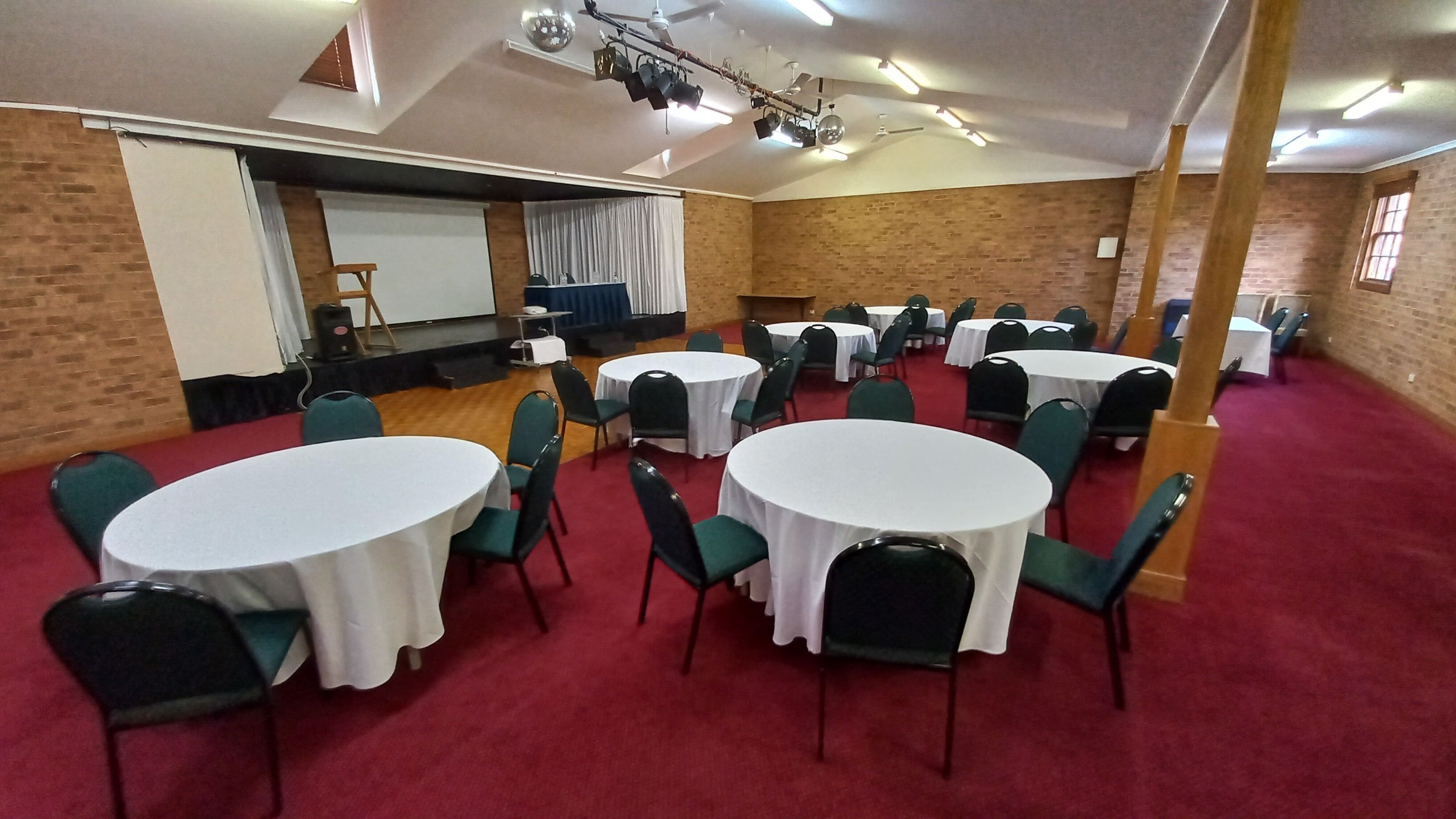 Un salón de conferencias con varias mesas redondas y sillas, un escenario con un proyector y una pared de ladrillo.