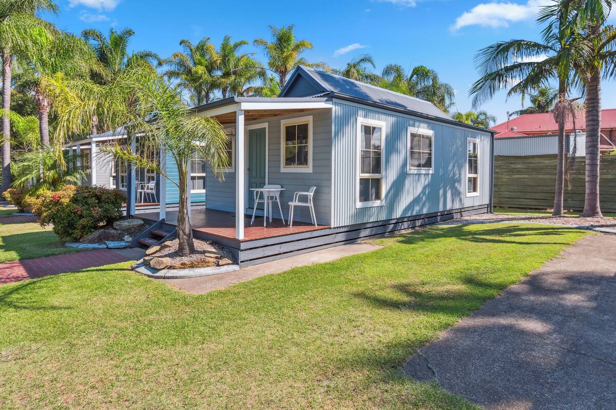 A small, single-story house with a porch, a small garden, and palm trees.
