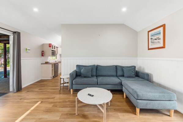 Una sala moderna con un sofá seccional gris, una mesa de centro redonda y una cocina al fondo.