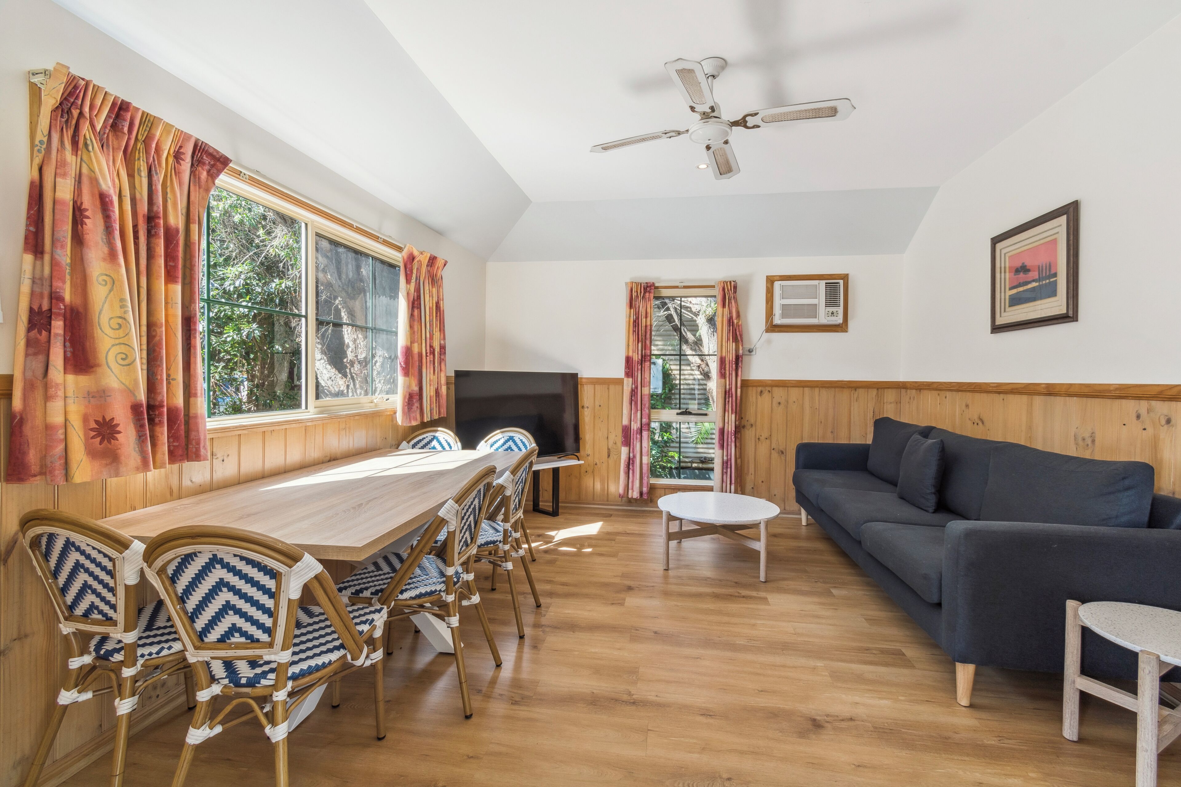 A room with a wooden dining table, chairs, a sofa, a television, and a ceiling fan.