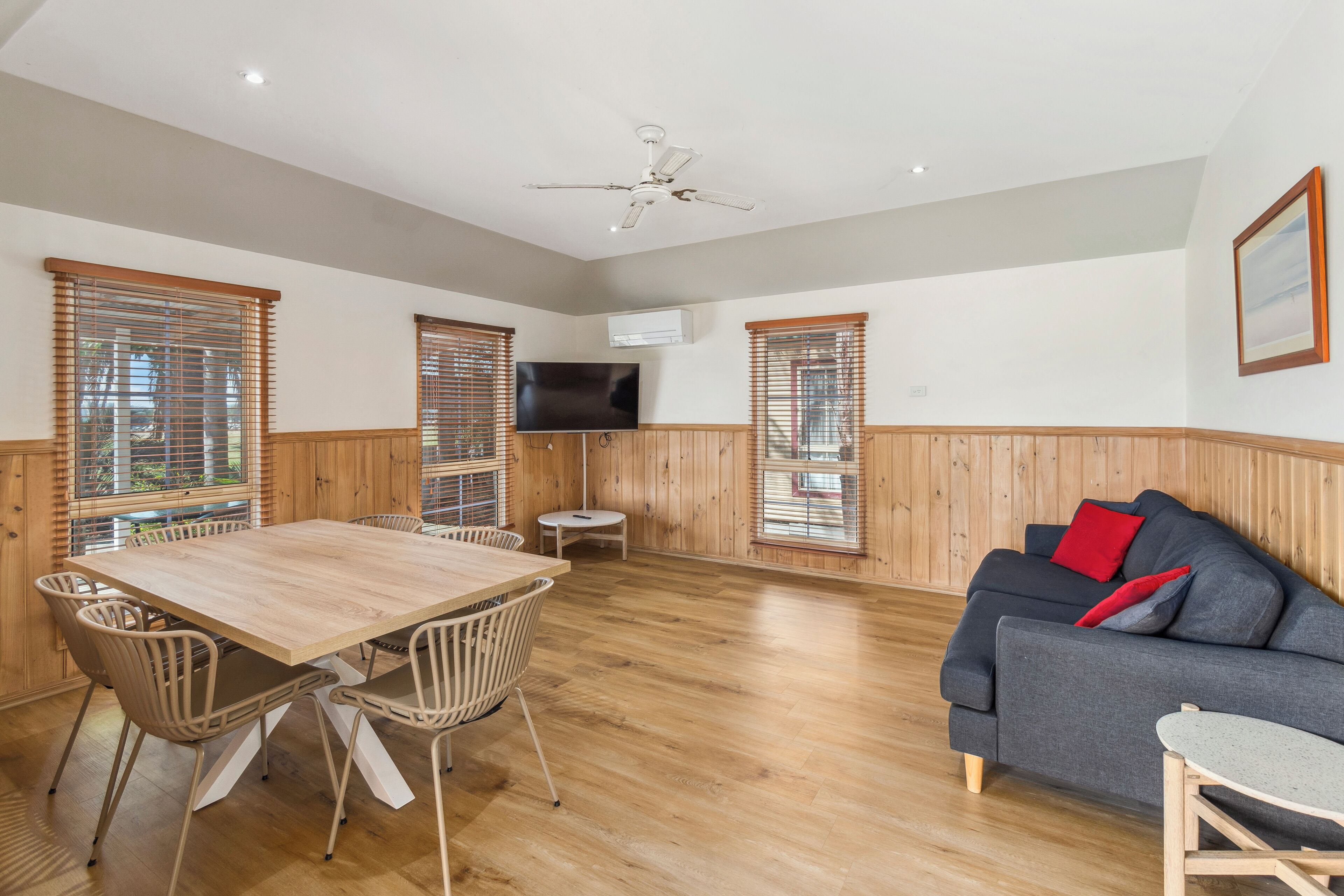 A spacious room with a wooden floor, a dining table with chairs, a grey sofa, and a ceiling fan.