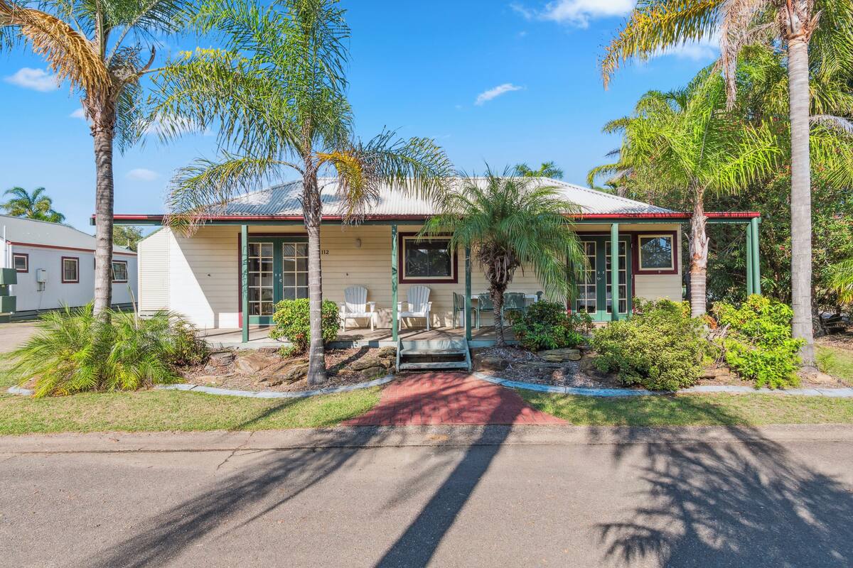 A single-story house with a red roof, white exterior walls, and a number '112' on the front. There are two white chairs outside, a small garden with palm trees, and a paved driveway.