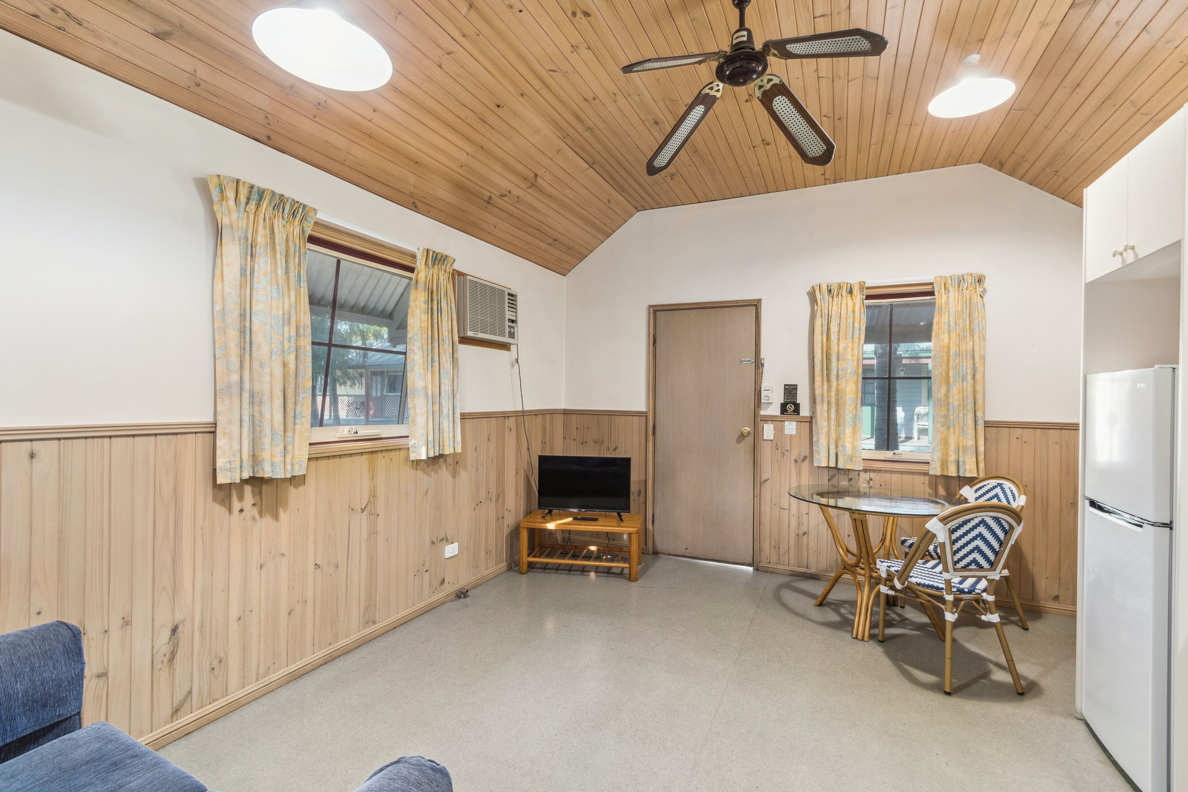 Une pièce aux murs et au plafond en bois, avec un ventilateur de plafond, un téléviseur posé sur un meuble en bois, une table à manger avec des chaises et un réfrigérateur.