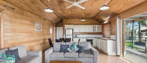 Un espacio habitable amplio con un conjunto de sofás y una mesa de centro, techo de madera con ventilador de techo, y una cocina al fondo.