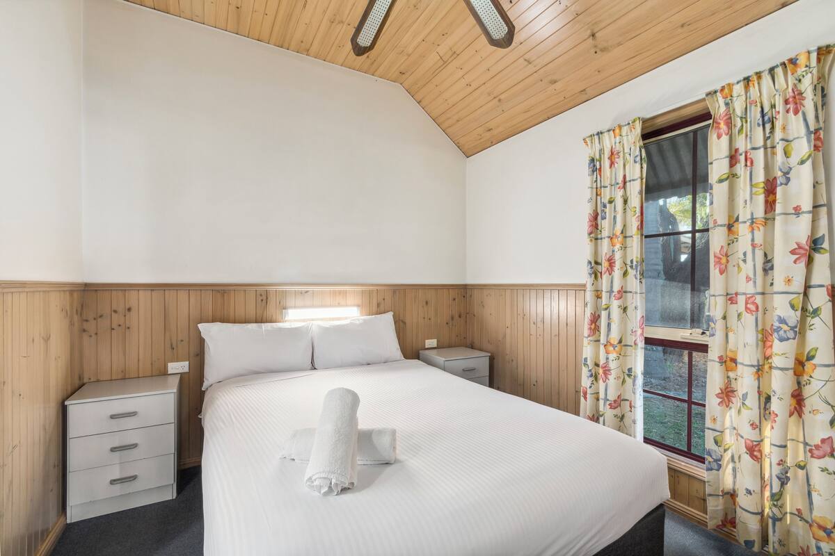 A bedroom with a wooden ceiling, a large bed with white bedding, a nightstand, a drawer unit, and a window with floral curtains.