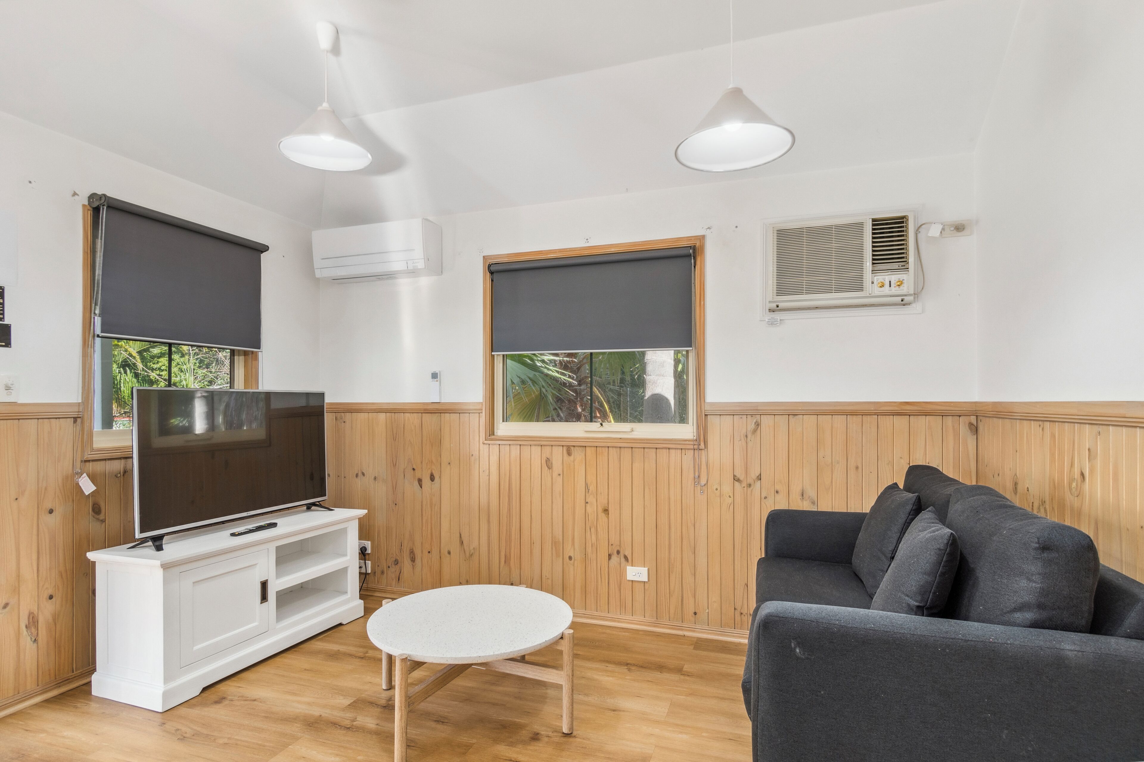 A living room with a TV on a white cabinet, a grey sofa, a round table, wooden paneled walls, and a window with a view of greenery.