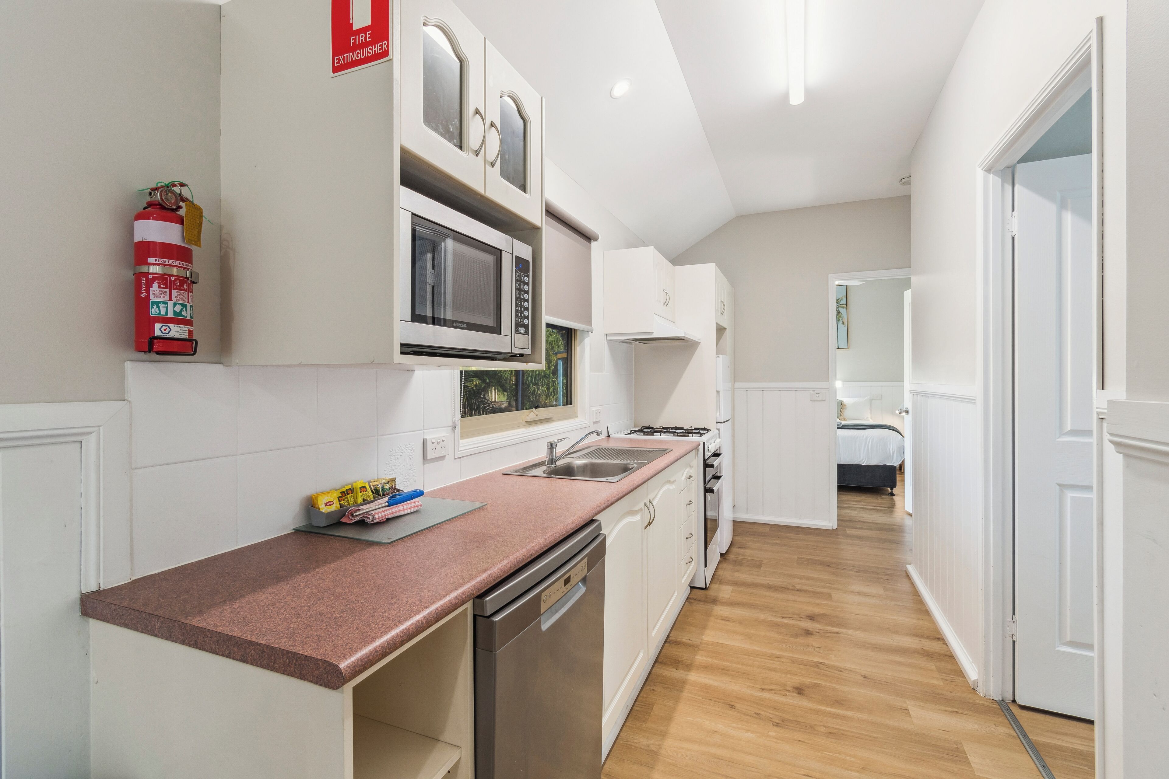 Cocina compacta con muebles blancos, lavavajillas de acero inoxidable, microondas y estufa a gas. Un extintor de incendios está montado en la pared.
