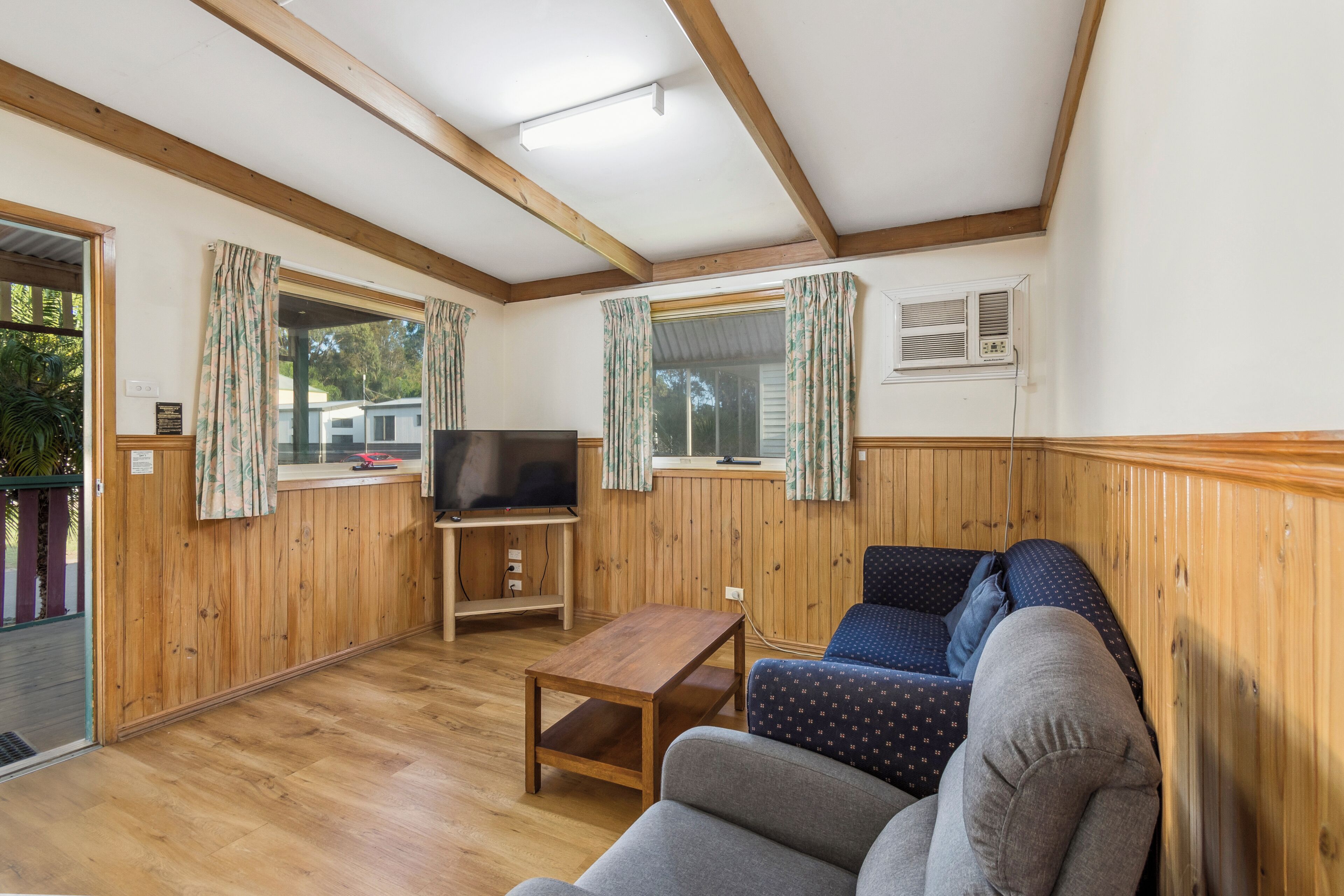 Quarto com paredes de madeira, televisão sobre suporte, sofá e janela com cortinas.