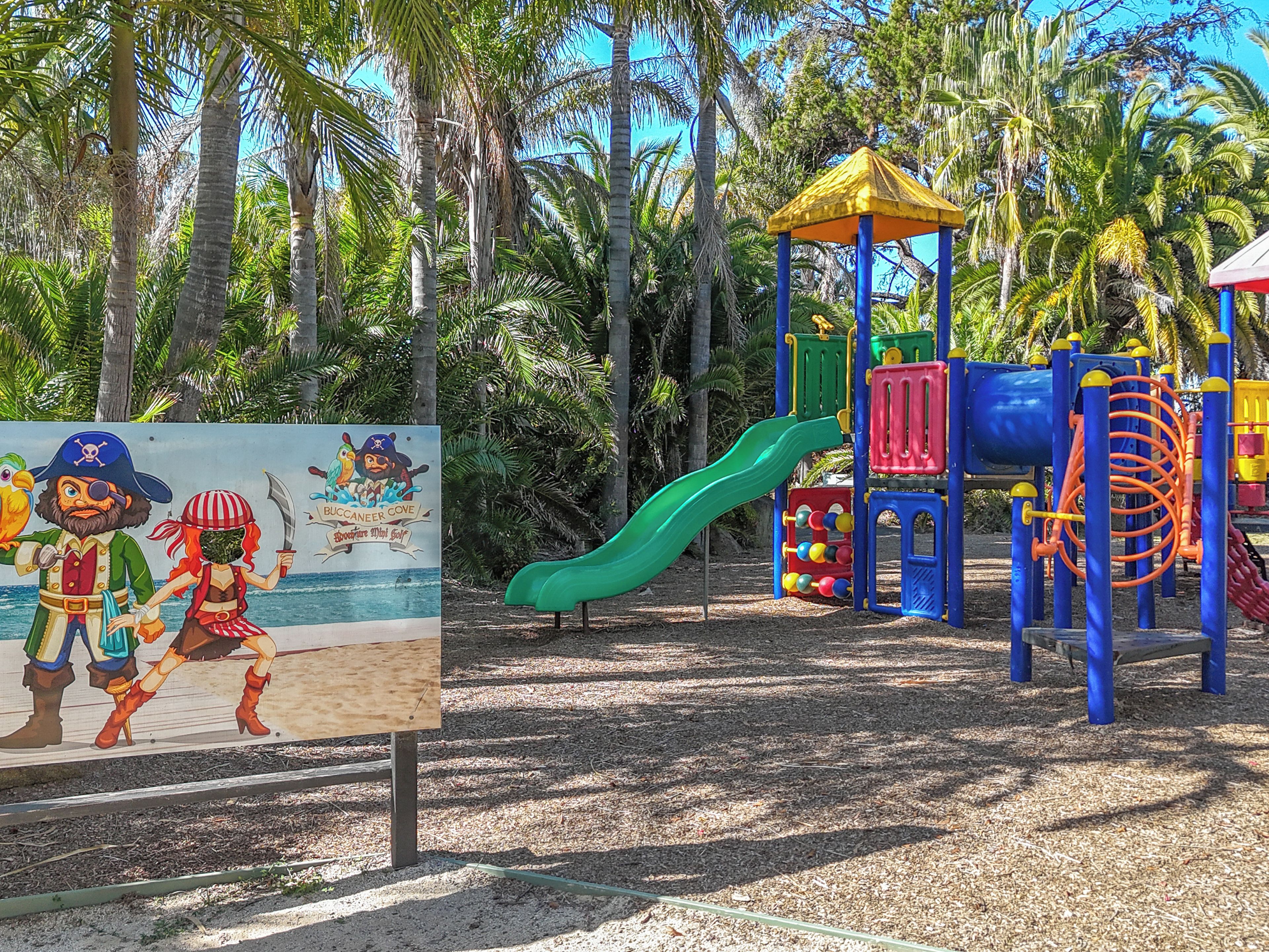 Un parque infantil con un tobogán y estructuras de escalada, rodeado de palmeras.