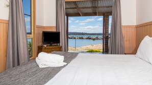 Habitación de hotel con una cama, un televisor sobre un soporte de madera, y vista a una playa y al agua a través de amplias ventanas.