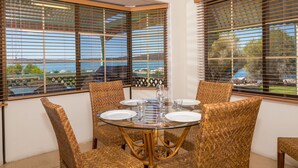 Un comedor con una mesa de vidrio rodeada de sillas de mimbre, ventanas amplias con persianas y vista al mar.