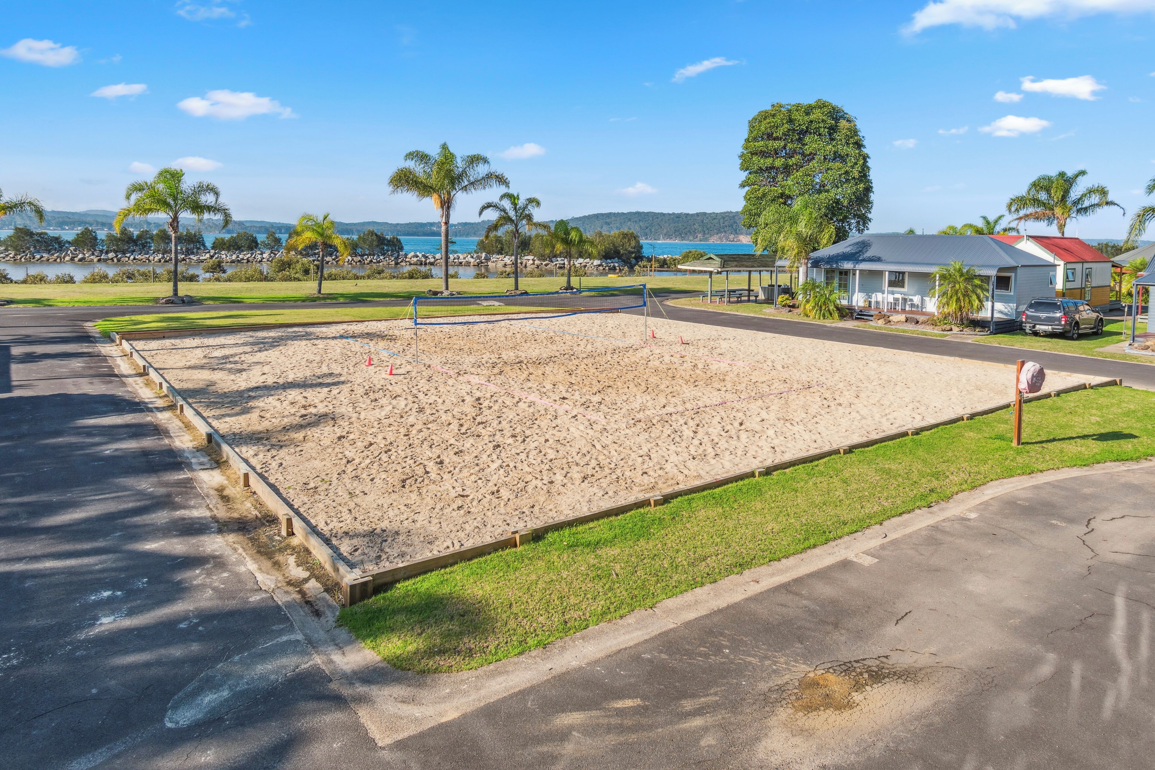 Una cancha de voleibol de playa con una red, rodeada de arena, césped y una carretera.