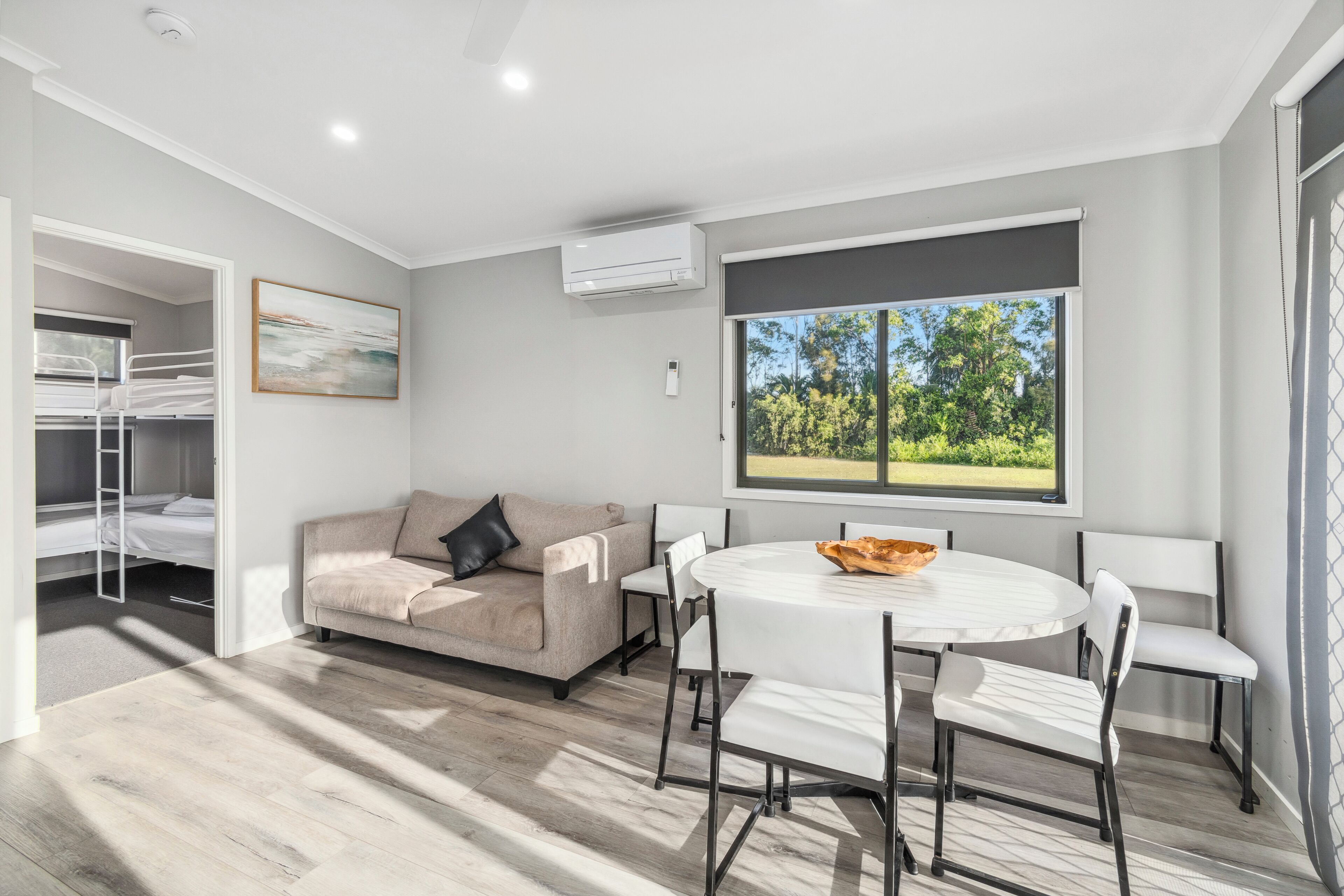 A modern living room with a beige sofa, a round dining table with white chairs, a window with a view of greenery, and an air conditioning unit on the wall.