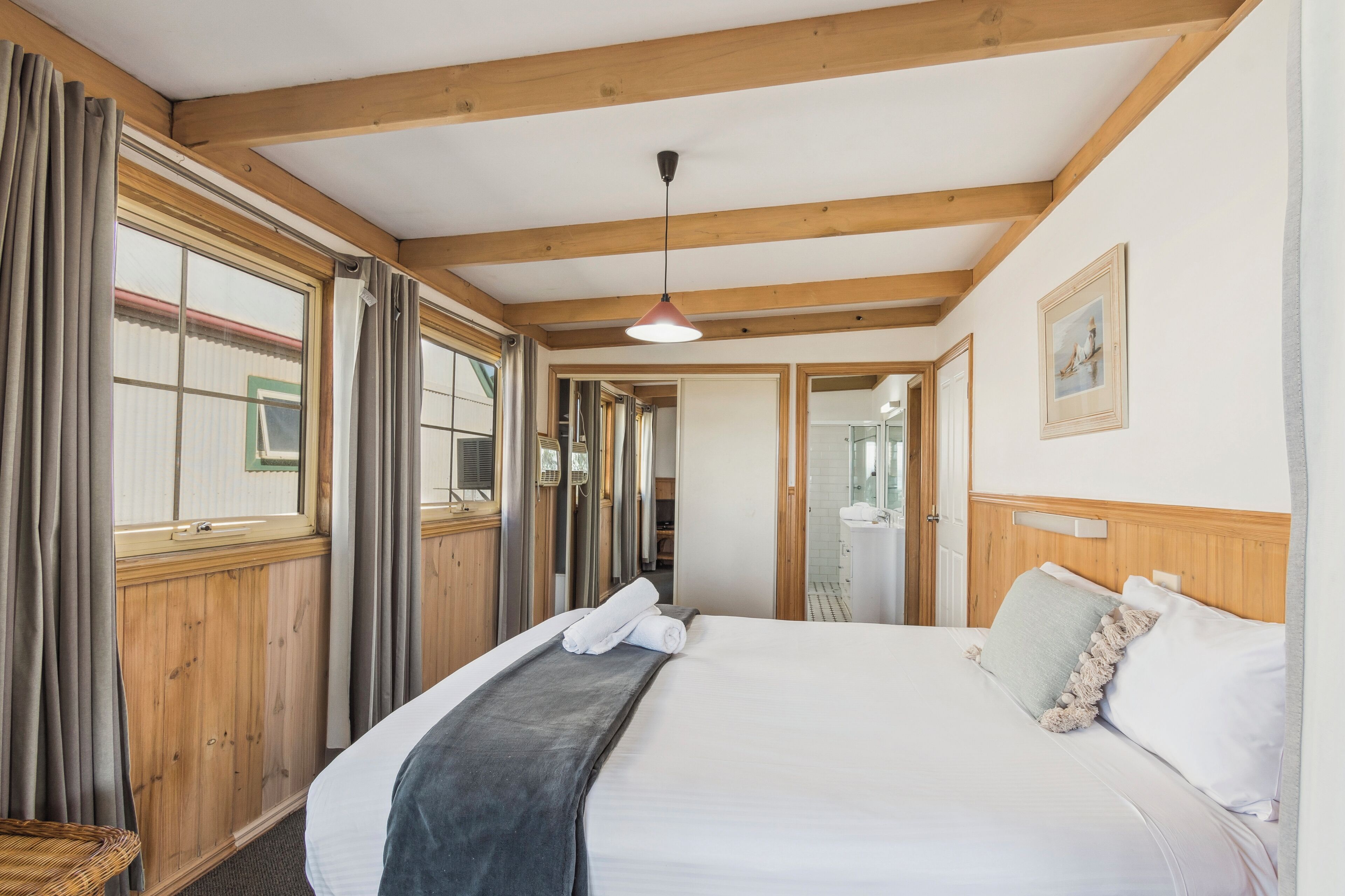 A hotel room with a large bed, wooden beams, a window with curtains, a bedside table, and a picture on the wall.