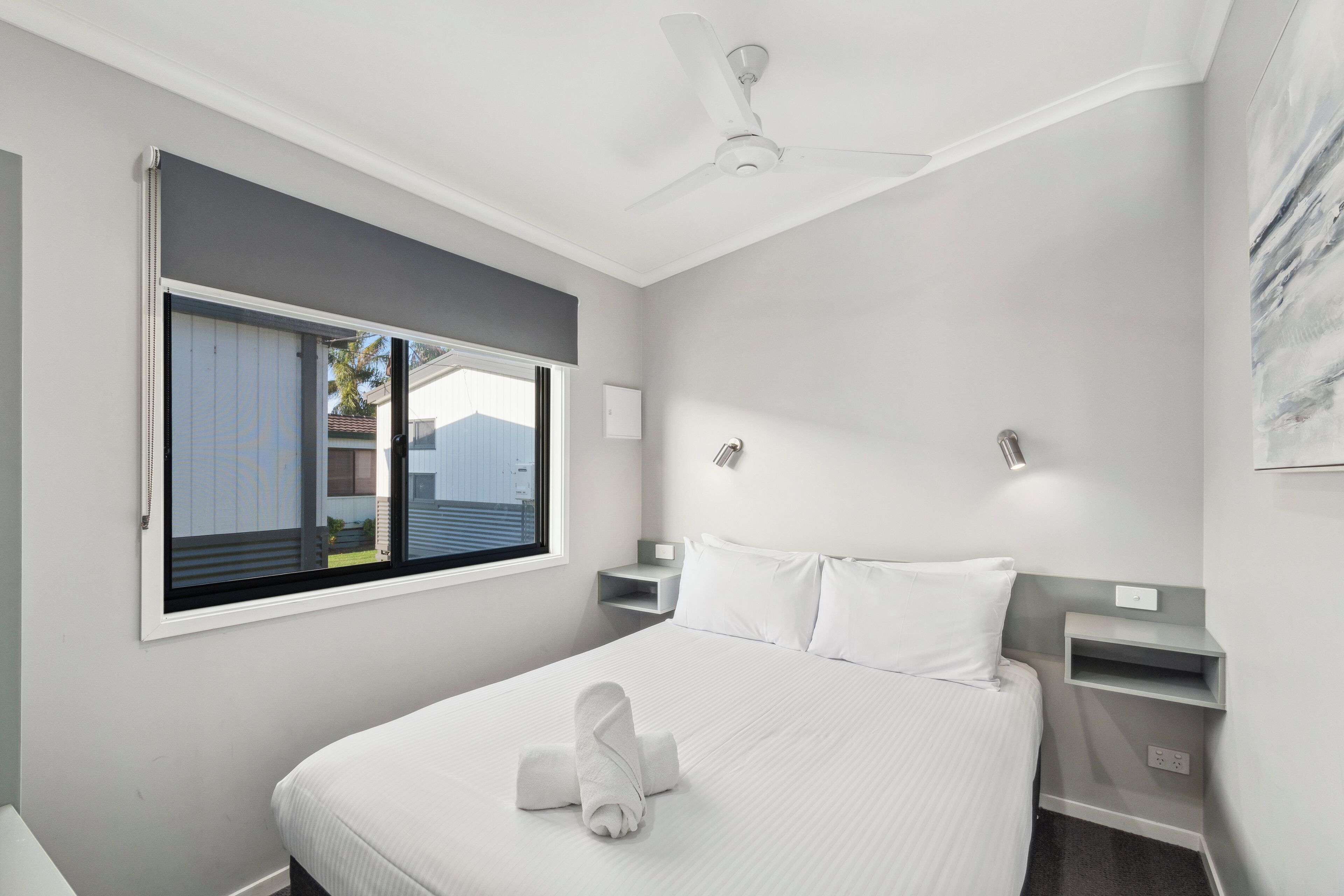 A modern hotel room with a large bed, a ceiling fan, a window with blinds, and a small bedside table with a lamp.