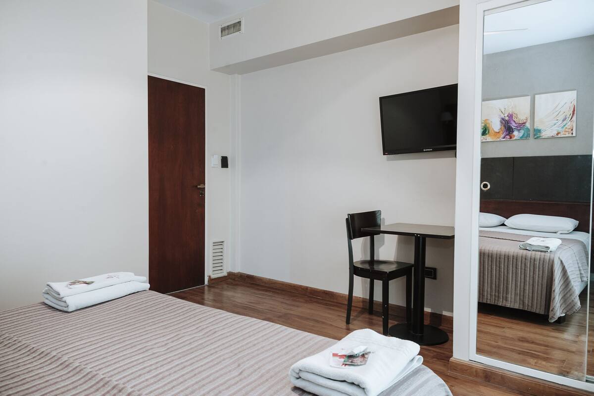 A hotel room with a bed, a desk, a chair, a television, and a mirror reflecting the bed and a painting.