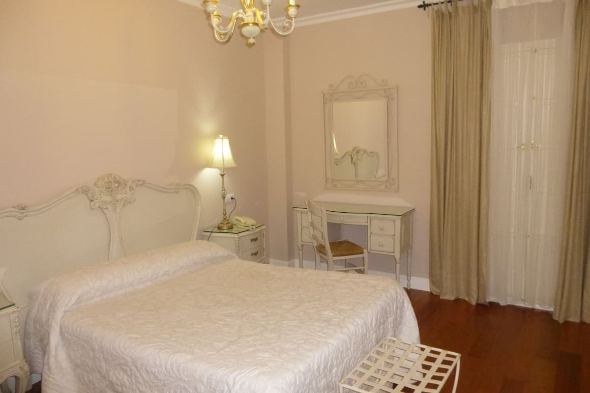Un dormitorio con una cama grande, una mesita de noche con lámpara, un aparador, una silla y un espejo.